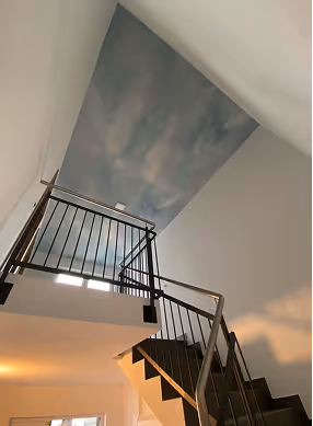 Treppe mit schwarzen Geländern und einer Decke mit Wolkenmalerei in einem hellen Innenraum.