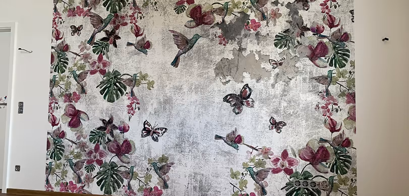 Wand mit Tapete, die rosa Orchideen, grüne Blätter, kolibriartige Vögel und Schmetterlinge auf einem verwaschenen grauen Hintergrund zeigt.