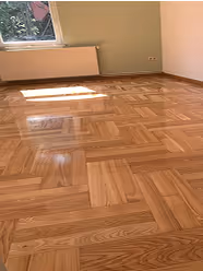 Leerer Raum mit einem hellen Holzboden im Fischgrätmuster, einer Heizung unter dem Fenster und beige Wänden.