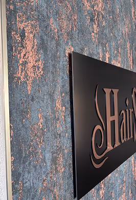 Seitliche Ansicht eines schwarzen Schilds mit dem Schriftzug 'Hair' an einer Wand mit rauer, dunkelblauer und kupferfarbener Textur.