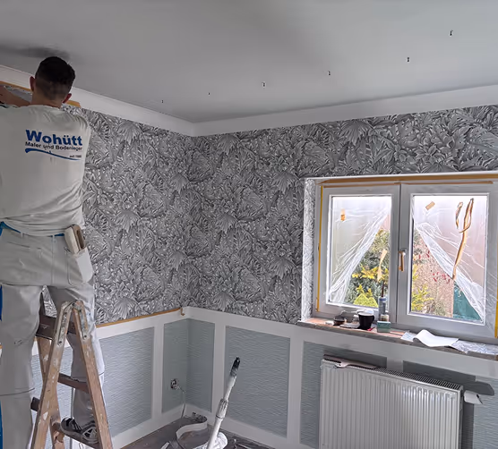 Maler steht auf einer Leiter und tapeziert eine Wand mit grauem, blättrigem Muster hinter einem Fenster, das mit Folie abgedeckt ist.