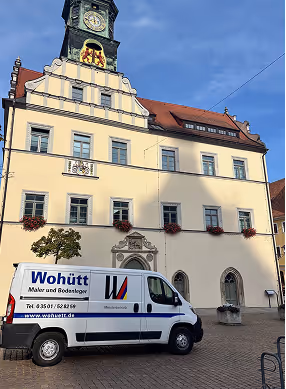 Weißer Kleintransporter mit Werbung für Maler und Bodenleger vor einem historischen Gebäude mit Turmuhr und blauem Himmel.