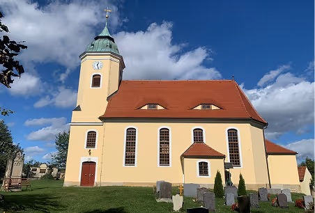 Gelbe Kirche mit rotem Dach und Uhrturm unter blauem Himmel mit weißen Wolken, davor ein Friedhof mit Grabsteinen.