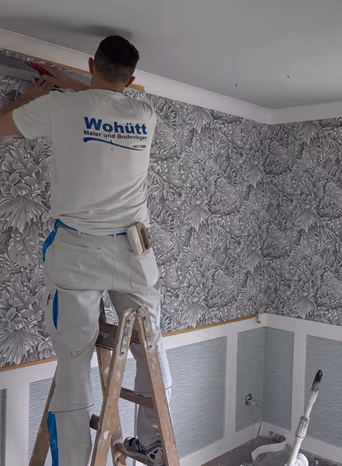 Handwerker steht auf einer Leiter und bringt Tapeten an einer Wand mit floralen Mustern an.